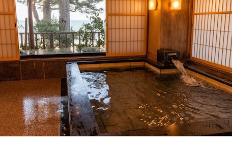 A traditional Japanese onsen with a wooden interior, a stone bath, and a view of trees and a wooden deck outside.