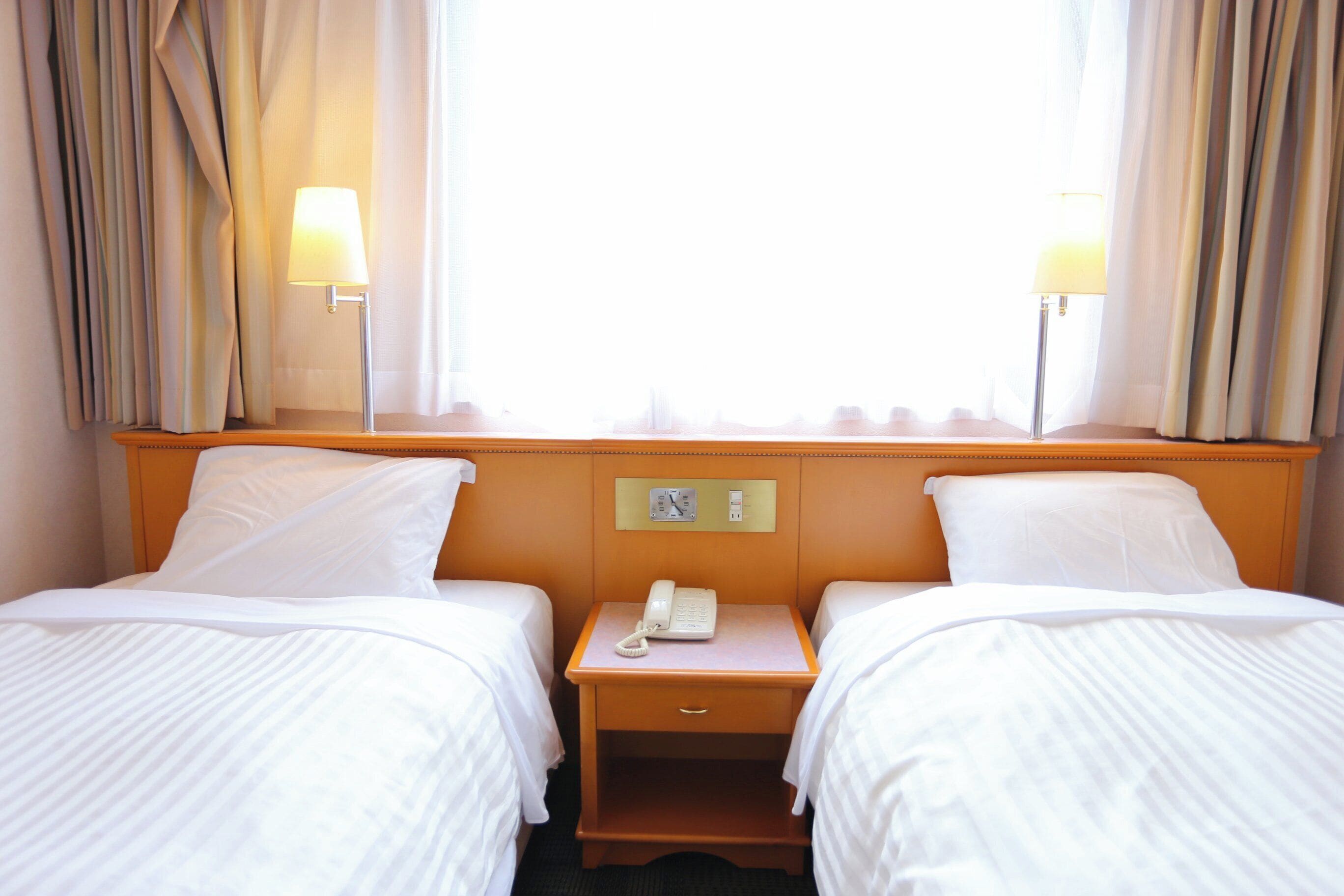 Een hotelkamer met twee bedden, een nachtkastje met een lamp, een telefoon en een raam met gordijnen.