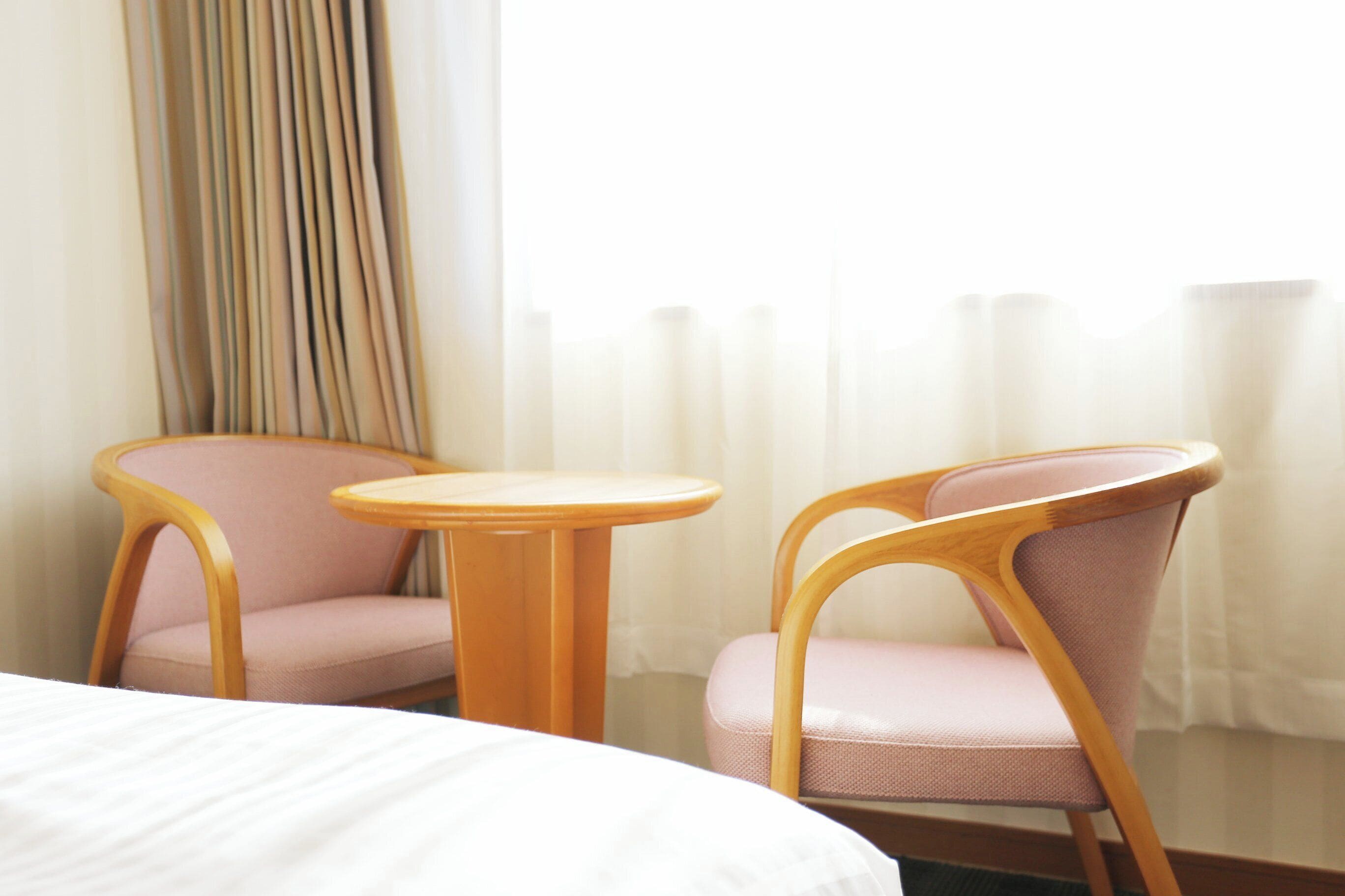 Habitación de hotel con una cama, dos sillas rosadas, una mesita de madera y una ventana con cortinas.
