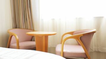 Une chambre d’hôtel avec un lit, deux fauteuils roses, une petite table en bois et une fenêtre avec des rideaux.