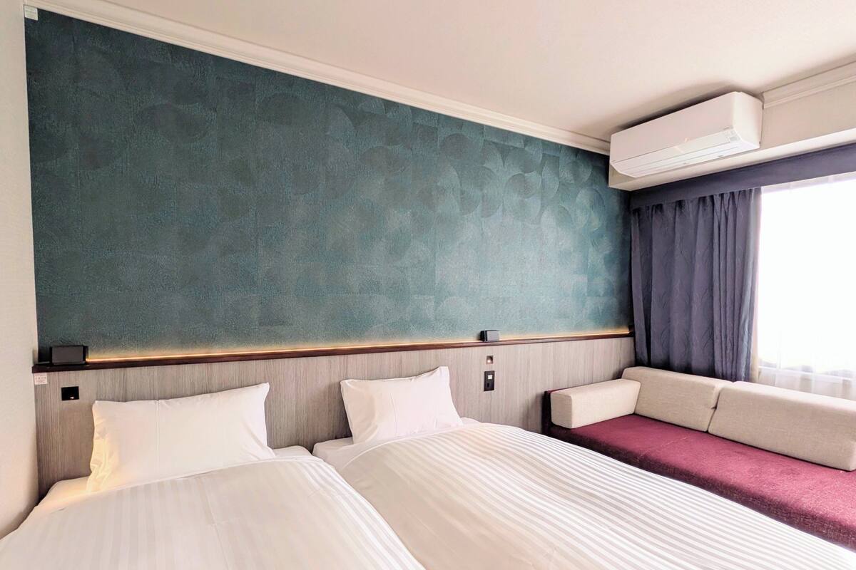 Habitación de hotel con dos camas, una pared verde texturizada, un equipo de aire acondicionado y una ventana con cortinas.