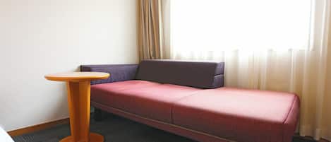 A room with a single bed, a small round table, a sofa, and a window with curtains.