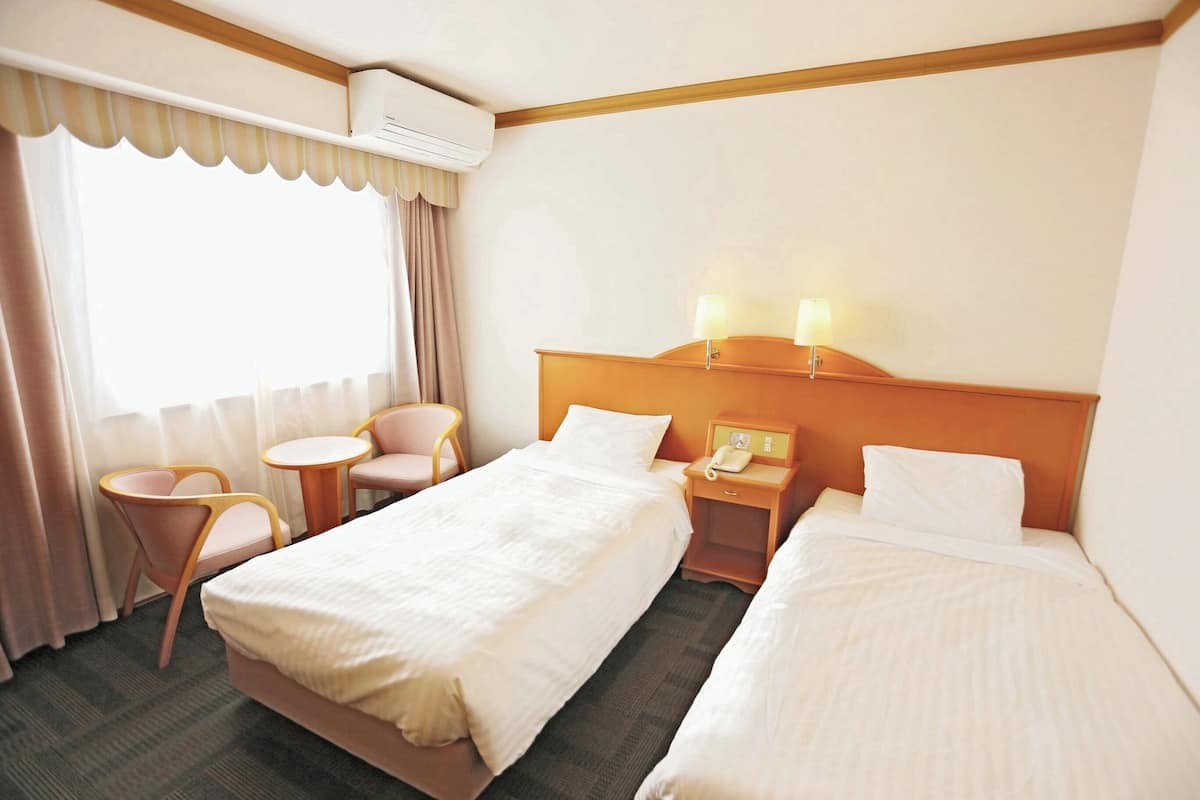 Una habitación de hotel con dos camas, una mesita, una silla, una mesita de noche con lámpara y un equipo de aire acondicionado.