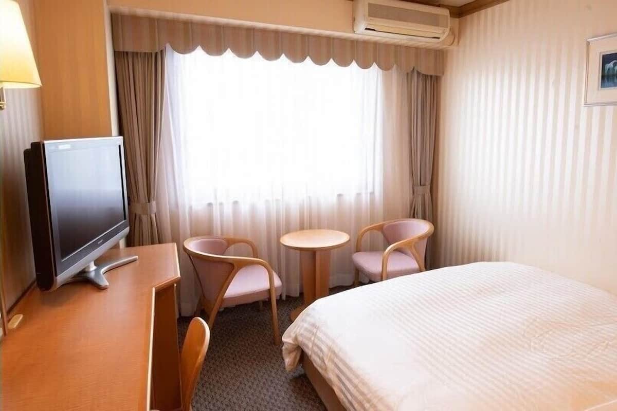 Habitación de hotel con cama, escritorio con televisor, dos sillas, una mesita y ventana con cortinas.
