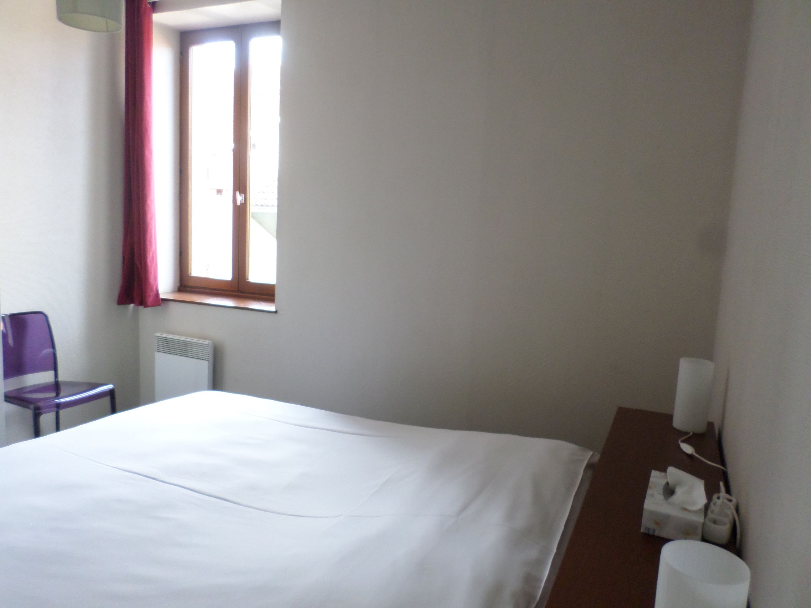 Een hotelkamer met een bed, een stoel, een raam met gordijnen, een radiator en een nachtkastje met zakdoekendoos en een lamp.