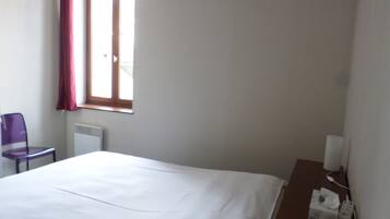 Een hotelkamer met een bed, een stoel, een raam met gordijnen, een radiator en een nachtkastje met zakdoekendoos en een lamp.