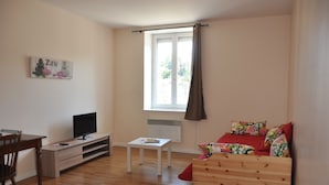 Ein kleiner, gut beleuchteter Raum mit Holzboden, einem weißen Schreibtisch mit Fernseher, einem kleinen Tisch, einem farbenfrohen Sofa und einem Fenster mit Vorhang.