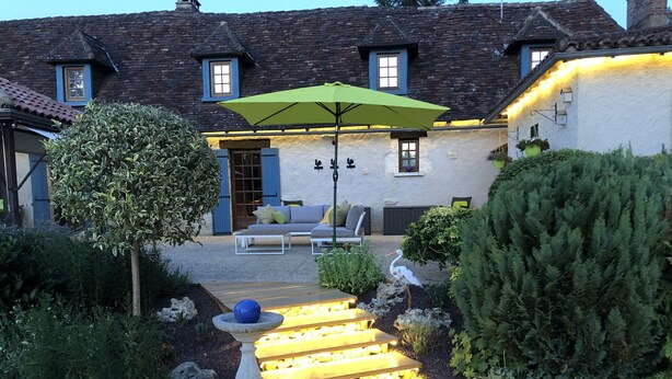 Un espace extérieur bien éclairé, aménagé avec des sièges, un parasol vert, un abreuvoir à oiseaux en pierre et un escalier en bois menant à une porte.