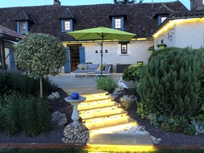 Un área de estar al aire libre bien iluminada, con un paraguas verde, un abrevadero de piedra y una escalera de madera que conduce a una puerta.