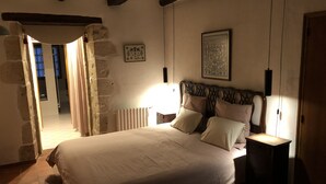 Une chambre avec un grand lit, un radiateur et une tête de lit en bois. On y trouve une porte qui donne sur une autre pièce et un tableau encadré accroché au mur.