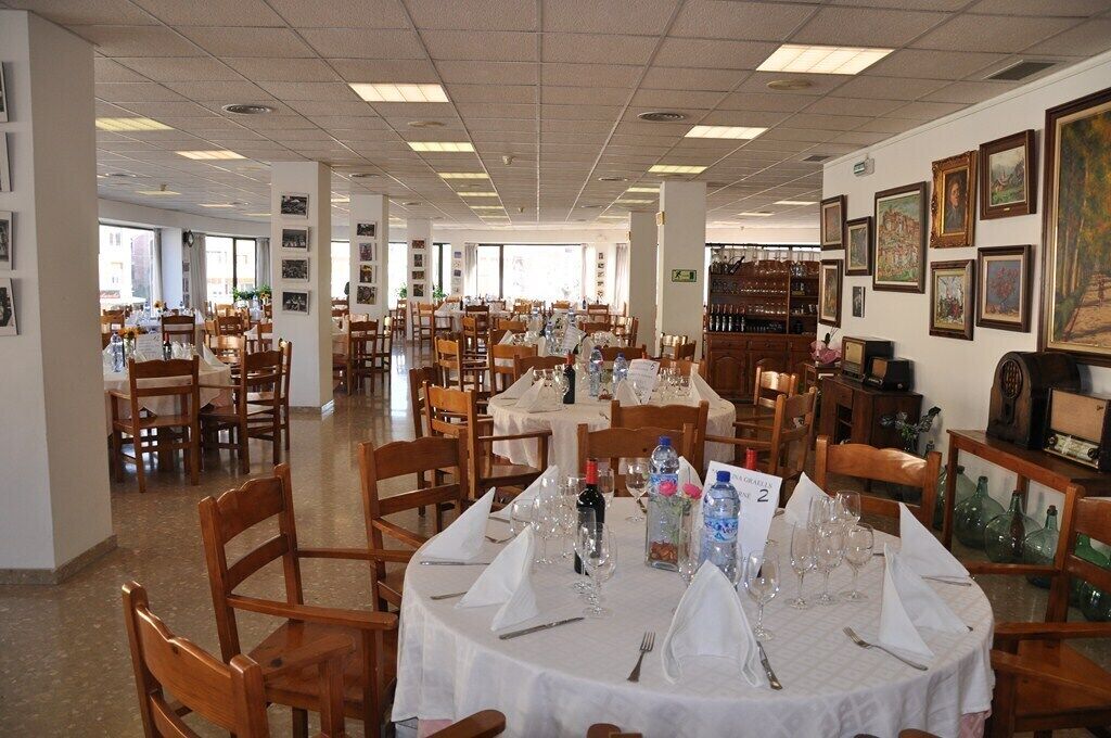 A spacious dining area with multiple round tables set for dining, wooden chairs, and a variety of table settings including wine glasses and water bottles.