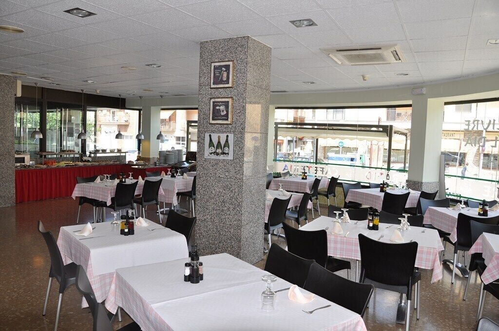 A restaurant with multiple tables, each covered with a white tablecloth and black chairs. There is a granite pillar with framed pictures and a menu board in the center.
