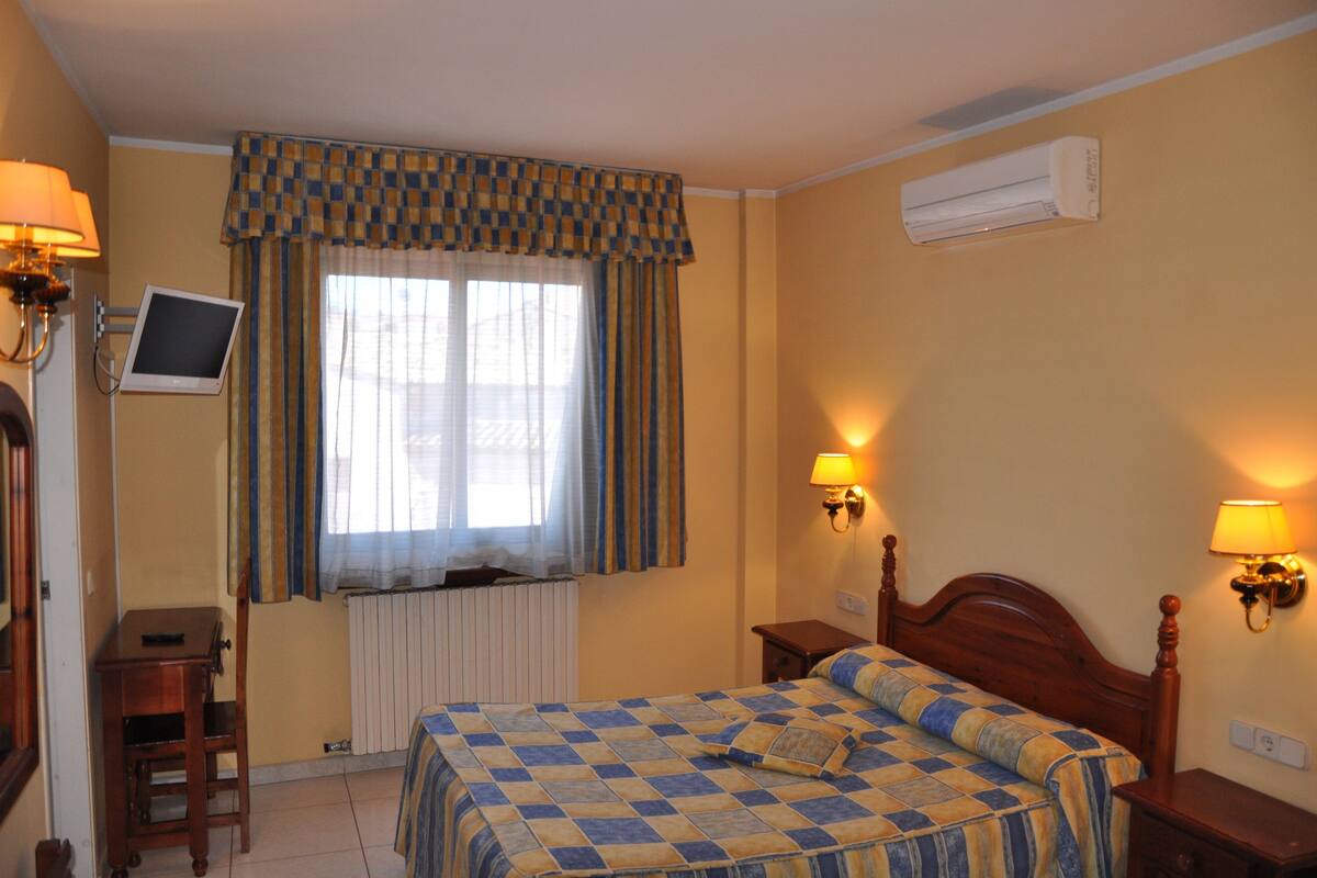 A hotel room with a checkered bedspread, a television on a stand, a window with curtains, and wall-mounted lamps.