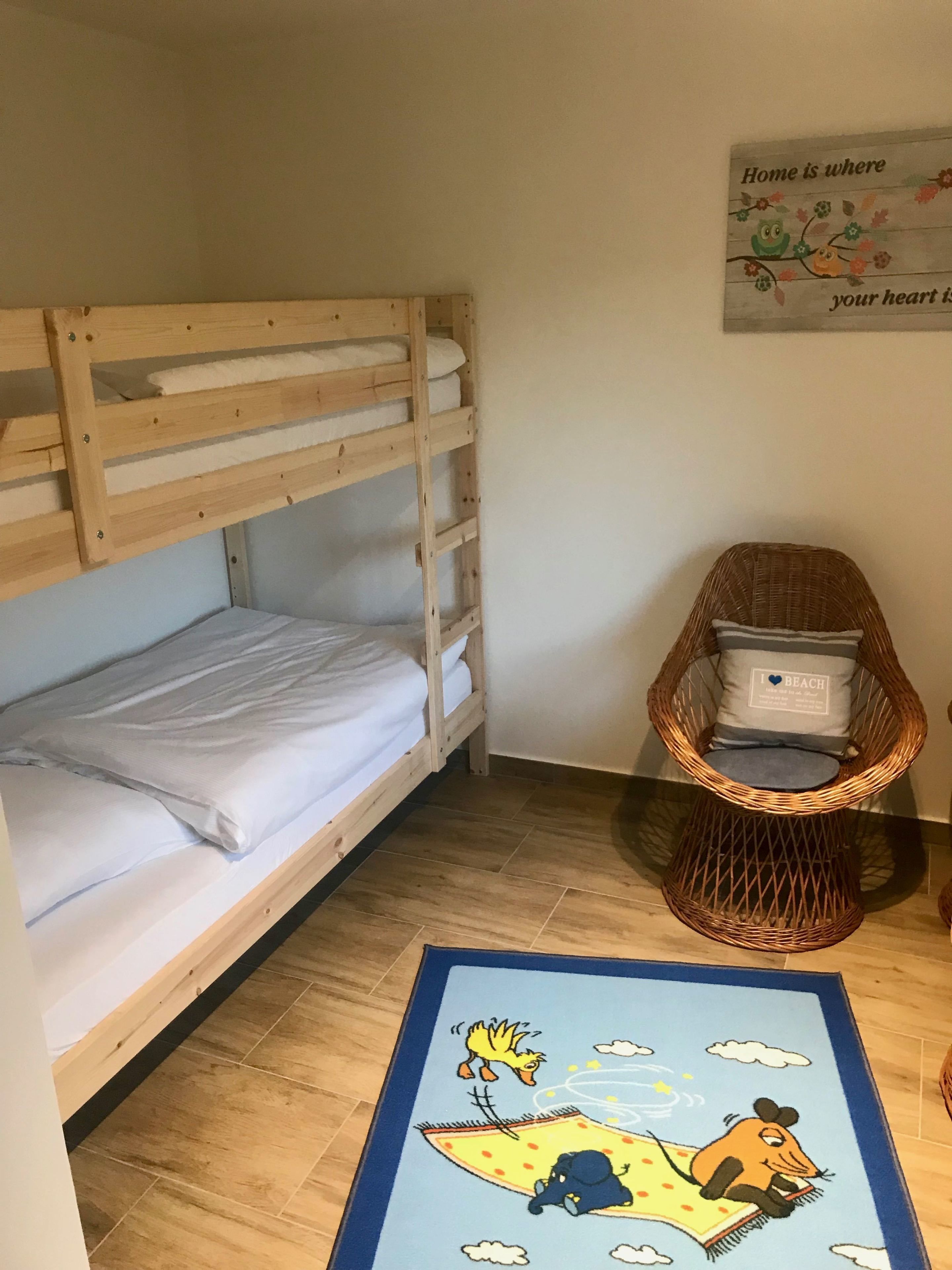 A bunk bed with white bedding, a wicker chair with a cushion, and a rug with a cartoon character.