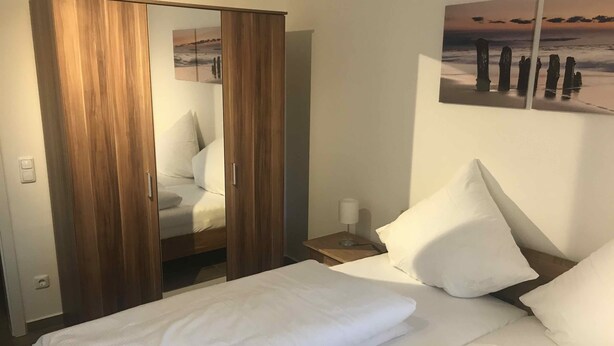 A hotel room with a bed, a wooden wardrobe, a nightstand with a lamp, and a wall-mounted painting of a beach scene. - Ferienwohnungen Möwe (Lembruch)
