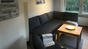 A modern living room with a dark grey sectional sofa, a small wooden table, and a window with a view of greenery. There is a wall sign with directions and a framed collage of nautical-themed photos. - Ferienwohnungen Möwe (Lembruch)