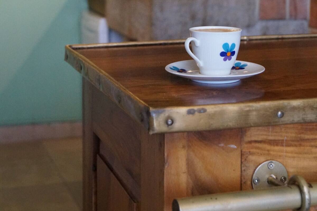 A wooden table with a white cup and saucer, a metal handle, and a brick wall with a tiled backsplash in the background.