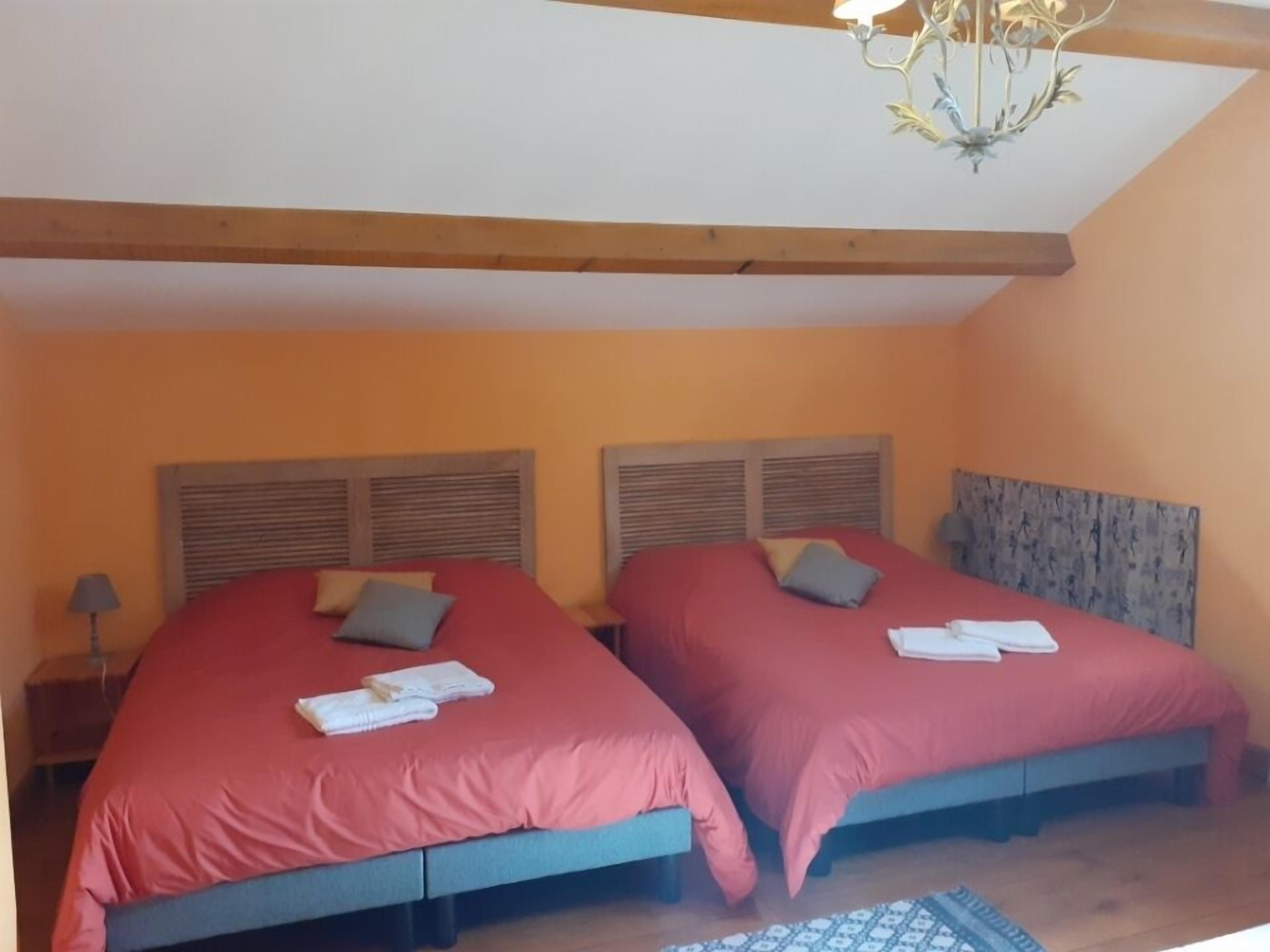 Un dormitorio con dos camas, cada una con cabecero de madera y ropa de cama roja. Hay una araña colgante sobre las camas y una mesita de noche con una lámpara a la izquierda.