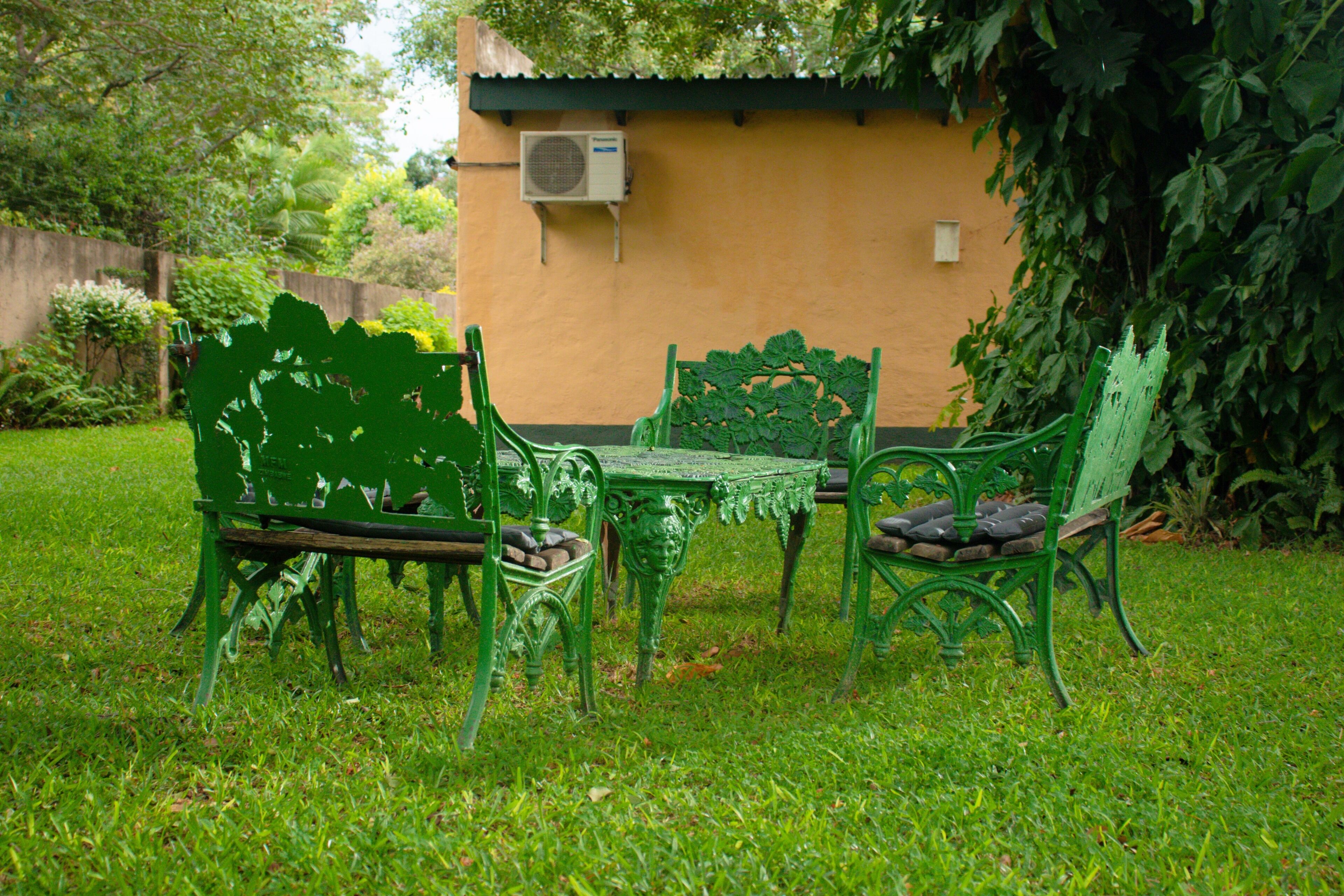 A green metal table and chairs set on a grassy lawn with a yellow wall and an air conditioning unit in the background.