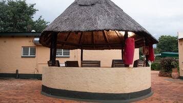 A thatched-roof gazebo with seating, a brick pathway, and a building in the background.