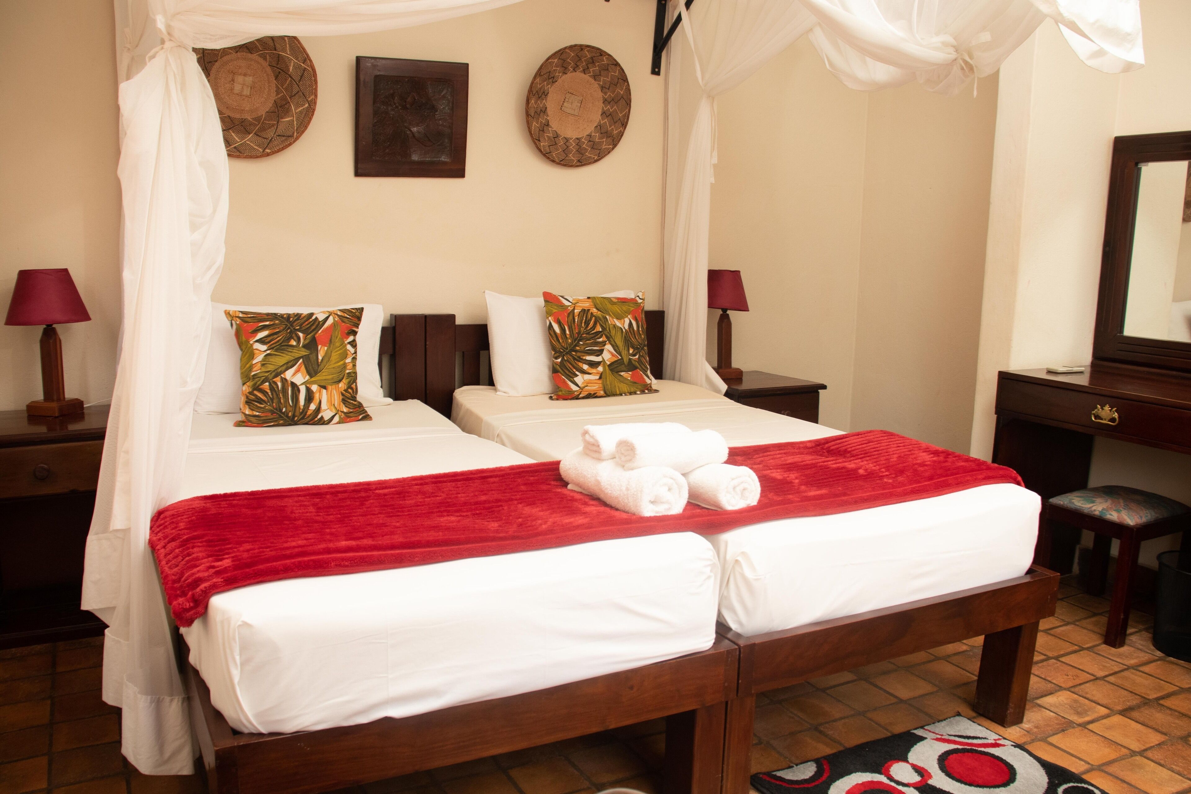 Un lit double avec du linge de lit blanc et une couverture rouge, deux oreillers ornés de motifs tropicaux, une table de chevet en bois avec une lampe et un petit tabouret.