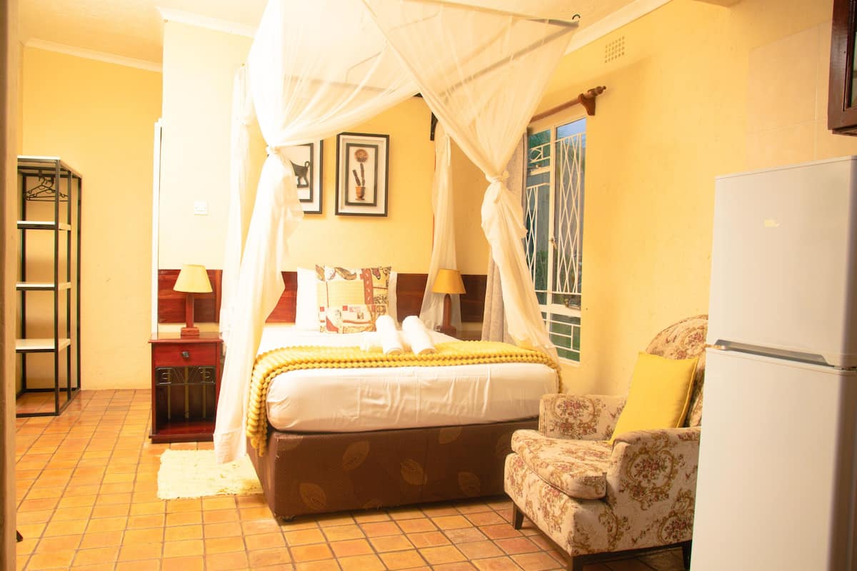 A bedroom with a bed, a chair, a bedside table, a lamp, and a refrigerator.