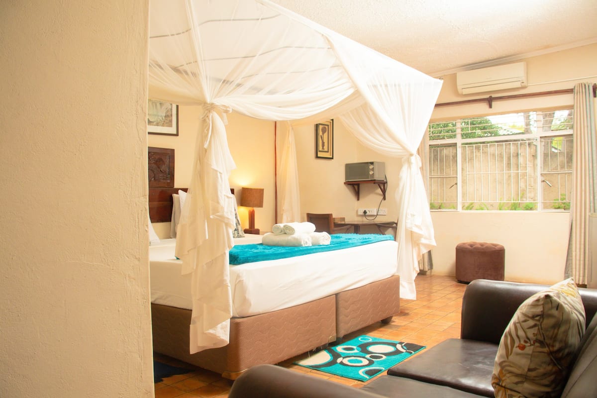 A bedroom with a four-poster bed, a desk with a chair, a laptop, and a window with a view of greenery.