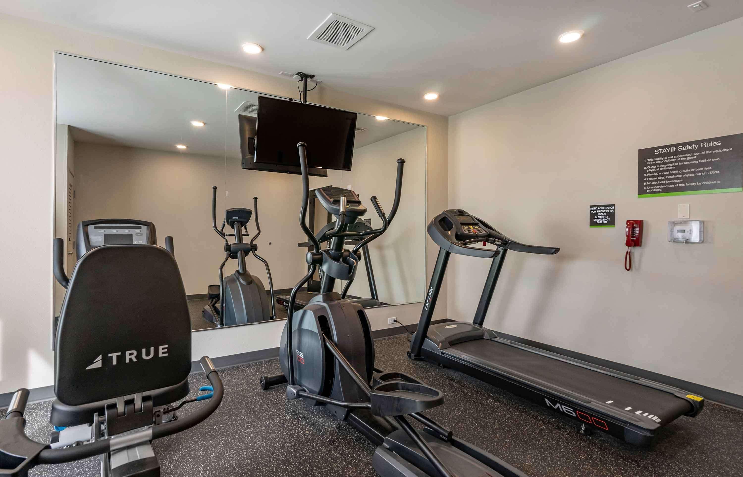 A gym with a treadmill, elliptical, and stationary bike, a TV mounted on the wall, and a sign with safety rules.