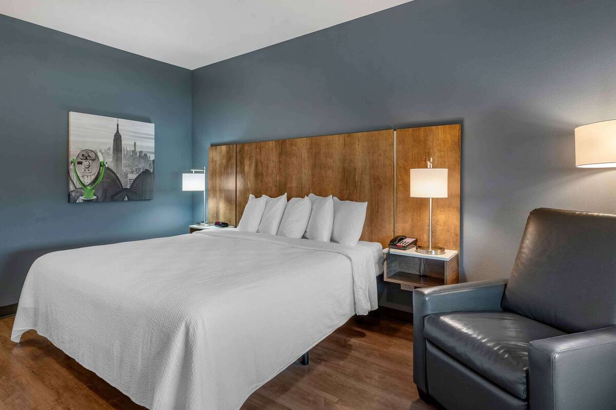 Ein Hotelzimmer mit einem großen Bett, einem Holzkopfteil, einem schwarzen Ledersessel, einem Nachttisch, einer Lampe und einem Bild an der Wand.