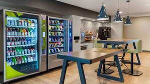 A modern hotel lobby with two beverage vending machines, a wooden table with chairs, and pendant lighting.