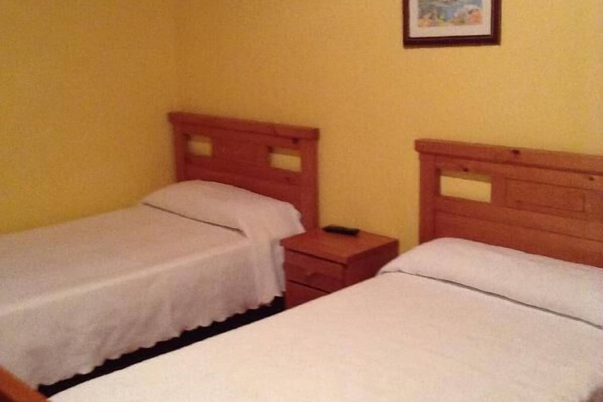 A room with two single beds, wooden headboards, a small bedside table with a remote control, and a framed picture on the wall.