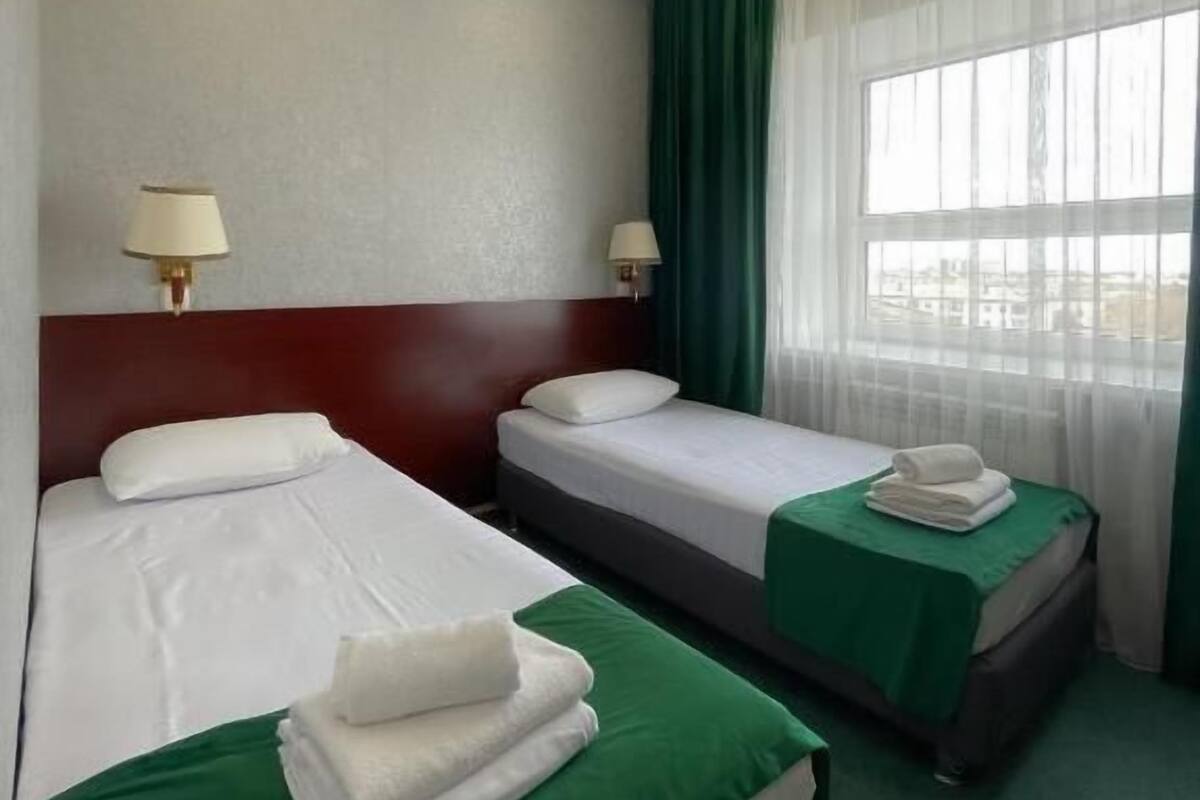 A hotel room with two beds, each with a green bedspread and white linens, a headboard, two bedside lamps, and a window with sheer curtains.