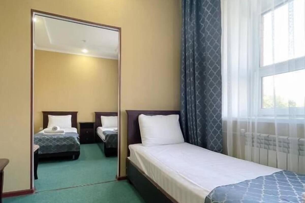 A hotel room with a single bed, a mirror, a window with curtains, and a green carpet.