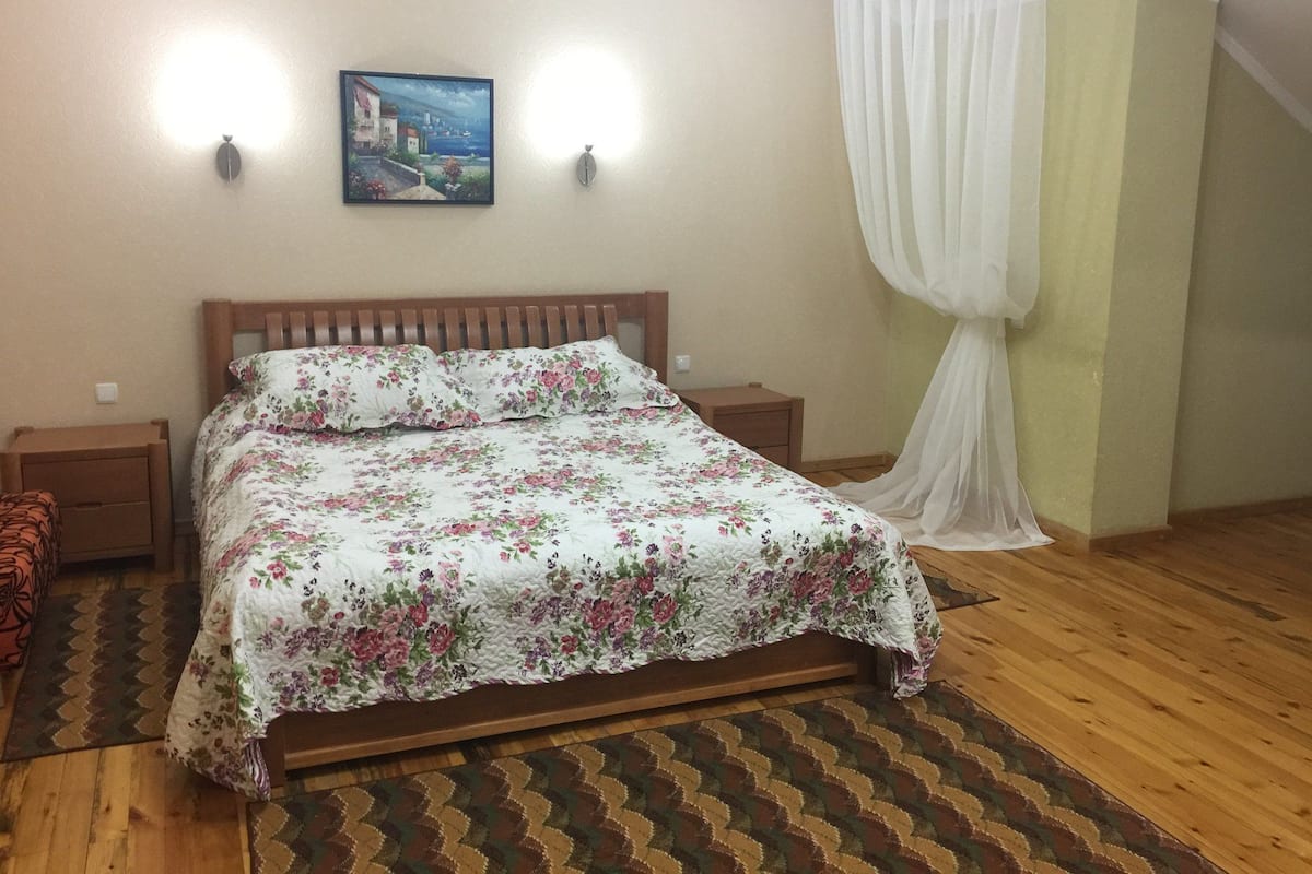 A bedroom with a wooden bed, floral bedding, two bedside tables, a nightstand, a painting on the wall, and a sheer curtain.