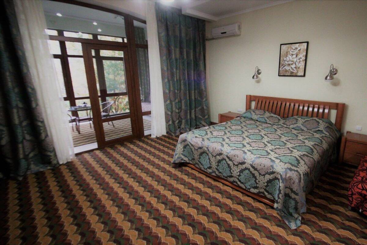 A bedroom with a patterned carpet, a bed with a patterned bedspread, a wooden headboard, two bedside tables, and a window with curtains.