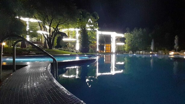 En nattöppen pool med ett kaklat poolområde, omgiven av träd och byggnader med upplysta fönster.