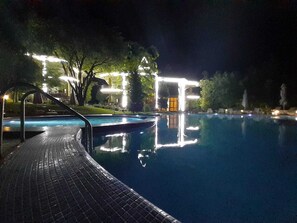A nighttime swimming pool with a tiled deck, surrounded by trees and buildings with illuminated windows. - Green Canyon Uzbekistan (Yusufhona)