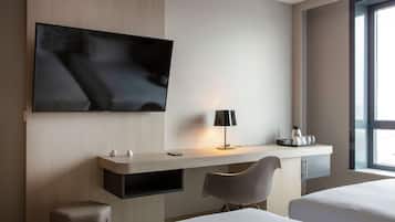 Ein modernes Hotelzimmer mit einem großen Flachbildfernseher an der Wand, einem Schreibtisch mit Lampe, einem Sessel und einem Bett mit weißer Bettwäsche.