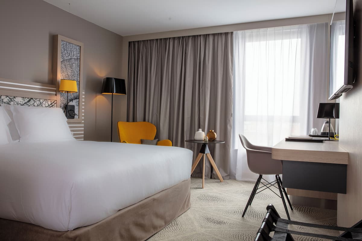 Ein Hotelzimmer mit einem großen Bett, einem Schreibtisch, zwei Stühlen, einem kleinen Tisch und einem großen Fenster mit Vorhängen.