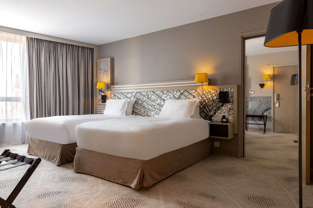 Ein Hotelzimmer mit zwei Betten, einem Kopfstück mit Muster, zwei Nachttischlampen, einem kleinen Tisch mit einer Uhr und Blick auf den Flur.