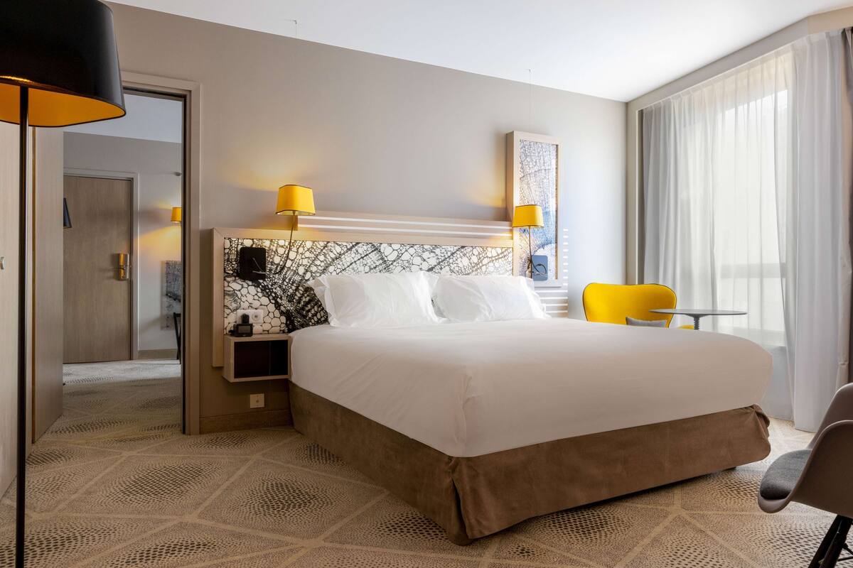 Ein Hotelzimmer mit einem großen Bett, zwei Nachttischlampen, einem kleinen Tisch, einem Sessel und einem großen Fenster mit Vorhängen.