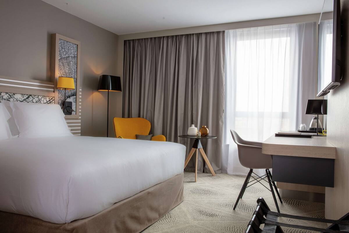 Ein Hotelzimmer mit einem großen Bett, einem Schreibtisch, einem Stuhl, einem kleinen Tisch und einem Fenster mit Vorhängen.