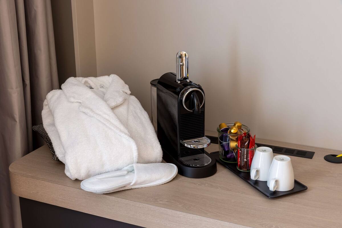 Ein Hotelzimmer mit einer Kaffeemaschine, einem Tablett mit zwei Tassen und Beilagen, einem weißen Bademantel und Hausschuhen auf einem Holztisch.
