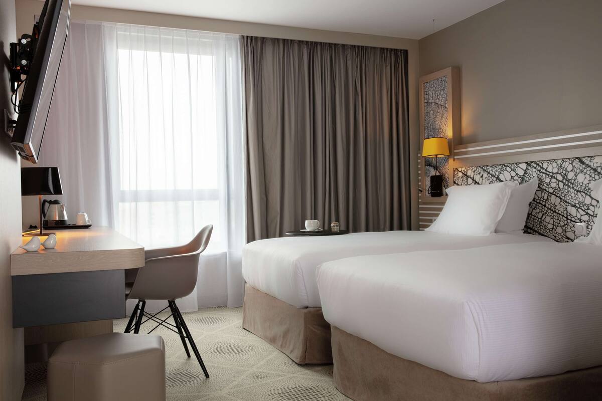 Ein Hotelzimmer mit Bett, Schreibtisch, Stuhl, Lampe und einem fenster mit Vorhängen.