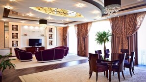 Una sala moderna con un televisor de pantalla plana, sofás morados oscuros, un comedor con mesa y sillas, y un ventanal con cortinas.
