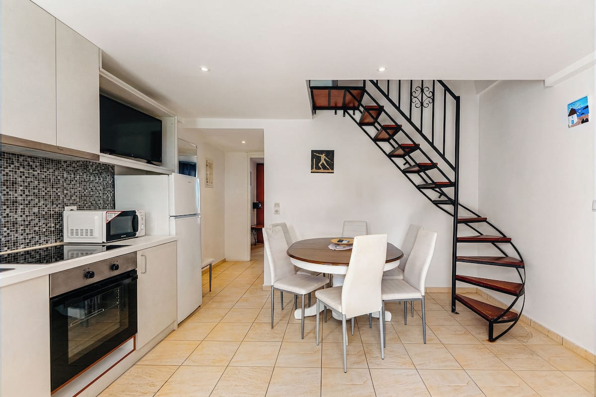 A modern kitchen with white cabinets, a microwave, and an oven. There's a dining table with chairs and a spiral staircase.