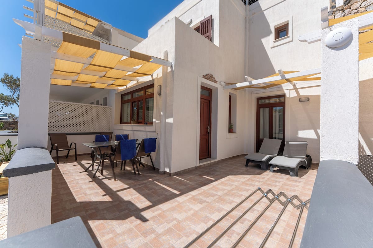 A covered outdoor area with a table and chairs, a poolside lounge, and a building with a white exterior and wooden accents.