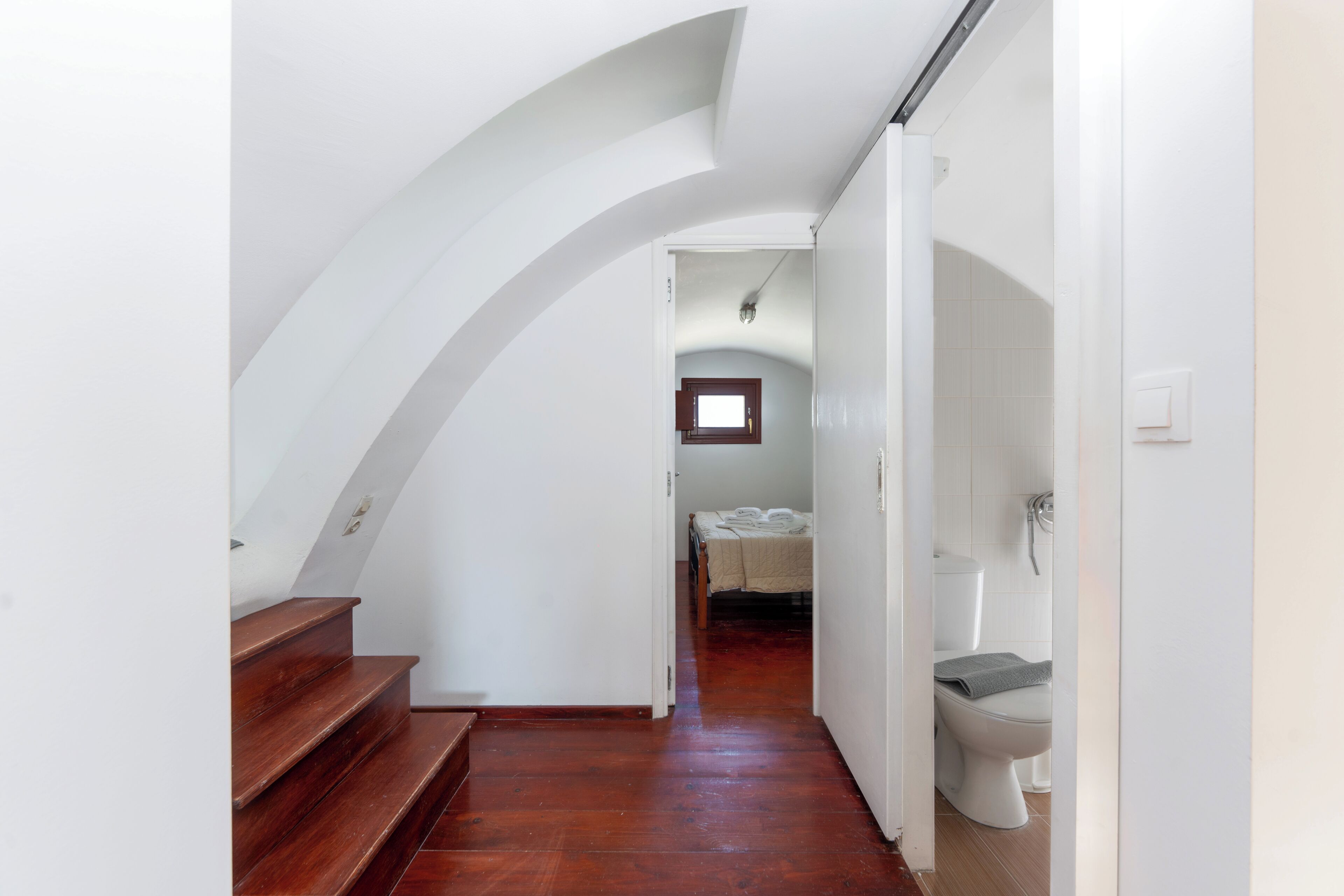 A narrow hallway with wooden stairs leading to a bedroom with a bed and a window.