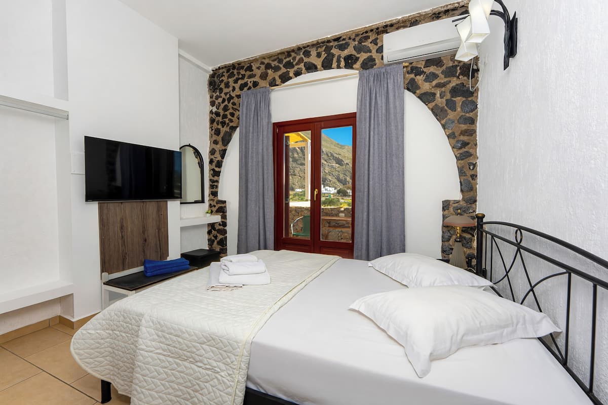 A bedroom with a bed, a flat-screen TV, a stone wall, and a view of the outdoors.