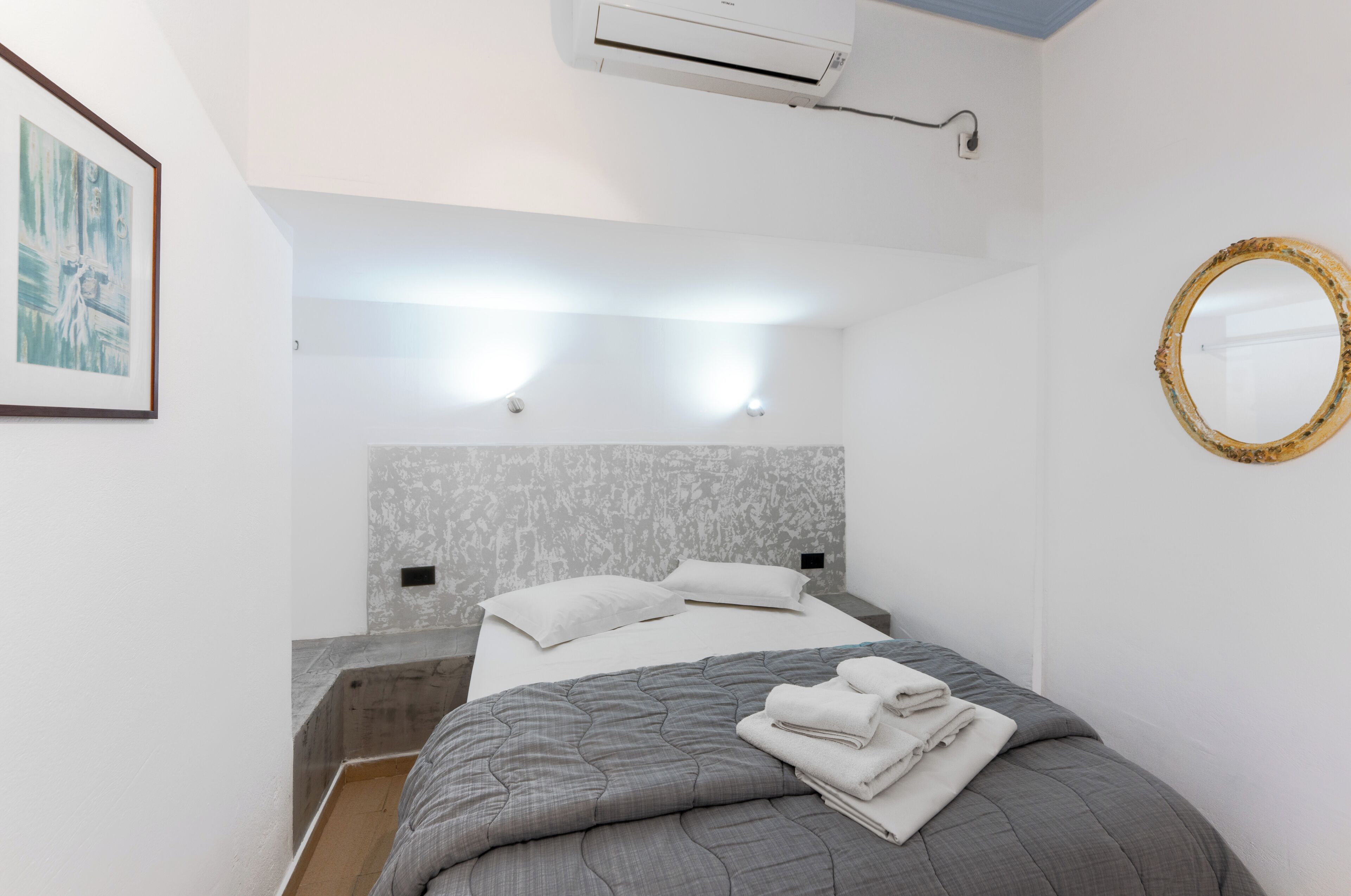 A modern hotel room with a bed, a headboard, an air conditioning unit, a wall-mounted mirror, and a framed picture.